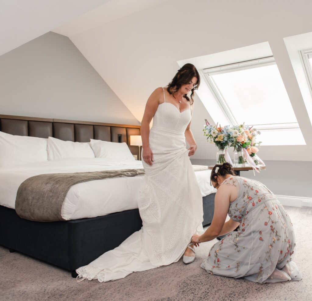 A bridesmaid helps dress the bride in our 5-star luxury bridal suite at Ness Walk Hotel In the Scottish Highlands for her wedding day.