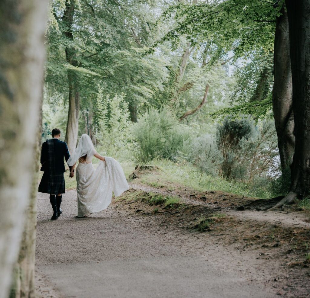 A Scottish wedding photo with a bride and groom walking along the banks of the River Ness with the groom in a kilt.