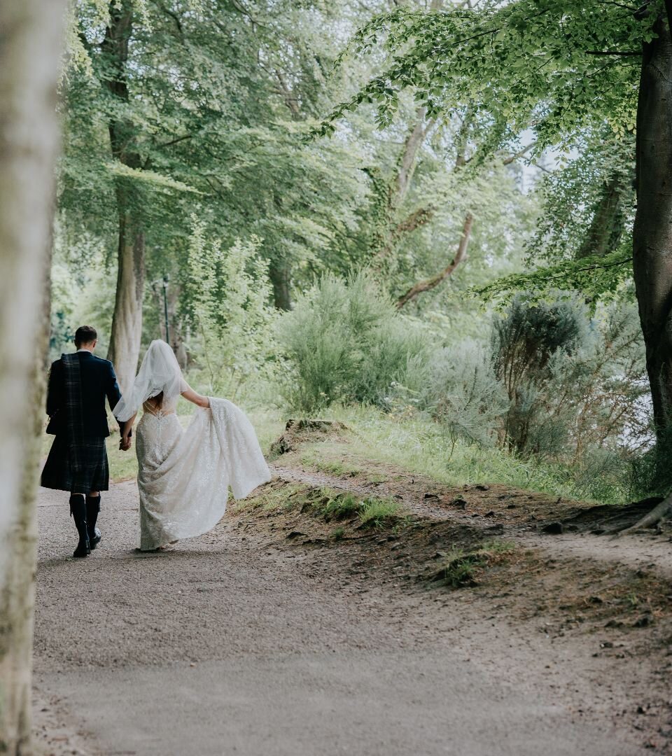 A Scottish wedding photo with a bride and groom walking along the banks of the River Ness with the groom in a kilt.
