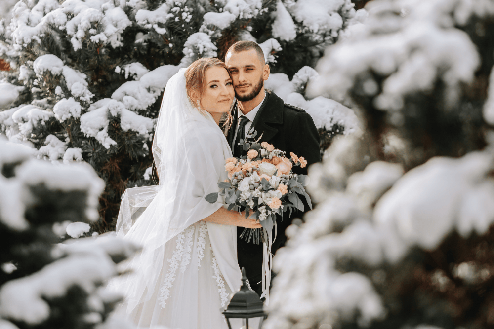 A couple embrace in wedding clothes in a snowy tree landscape.