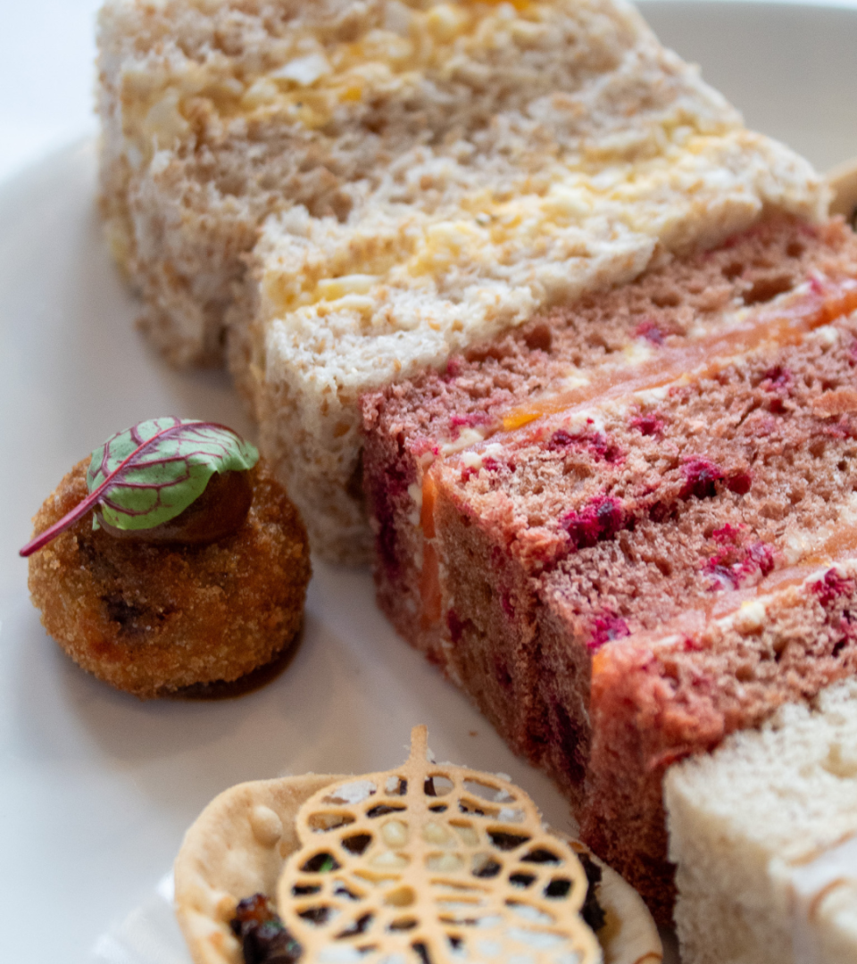 A close-up of elegant afternoon tea finger sandwiches on a white plate, including white bread egg mayonnaise and pink beetroot bread with smoked salmon, alongside a crumbed savoury ball garnished with a micro herb leaf and a decorative pastry tartlet with a lace-cut pastry lid