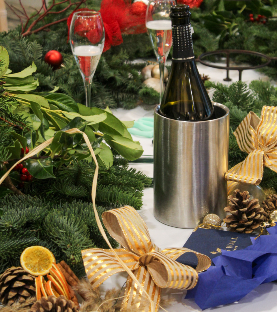 A Christmas workshop table display featuring a champagne bottle in a silver ice bucket, flute glasses, gold striped ribbon bows, pine cones, dried orange slices, cinnamon sticks, and lush evergreen foliage
