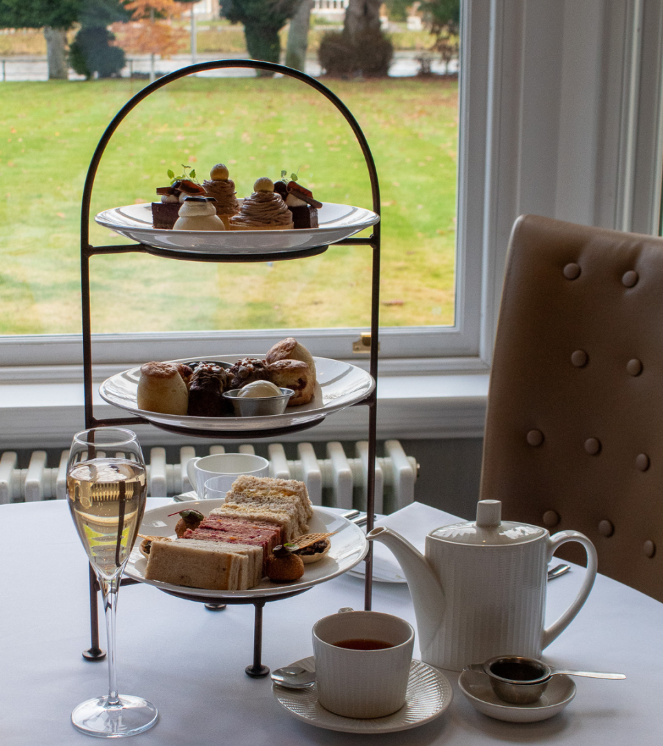 three-tiered afternoon tea stand by a large window overlooking a winter garden, set with finger sandwiches and savoury bites on the bottom tier, scones with accompaniments on the middle tier, and elegant patisserie on the top tier, alongside a glass of champagne, a white teapot, and a cup of tea