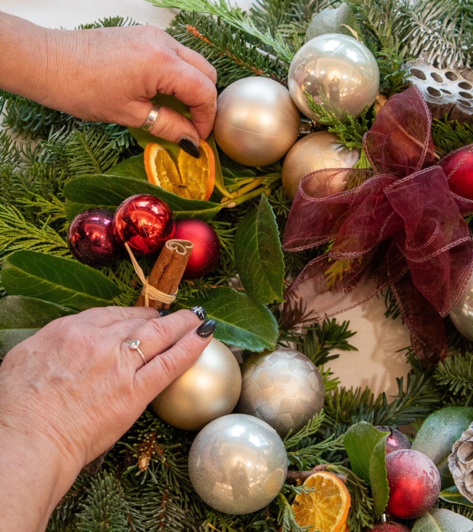 Close-up of a participant's hands arranging decorations on a Festive Table Arrangement including silver and red baubles, dried orange slices, cinnamon sticks, bay leaves, and a deep red ribbon bow