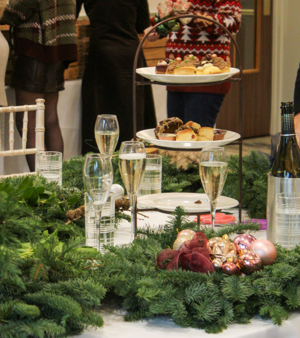 A Christmas workshop table set with champagne flutes, a three-tiered cake stand of festive bakes and pastries, evergreen garland, and rose gold and burgundy baubles, with guests mingling in the background