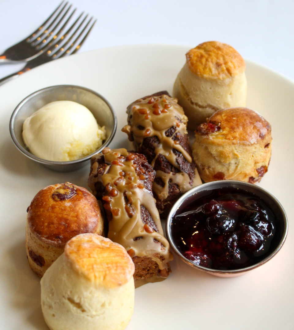 A plate of freshly baked plain and fruit scones alongside glazed chocolate and caramel tea loaf slices, served with a silver dish of clotted cream and a pot of dark berry jam, on a white plate with a fork