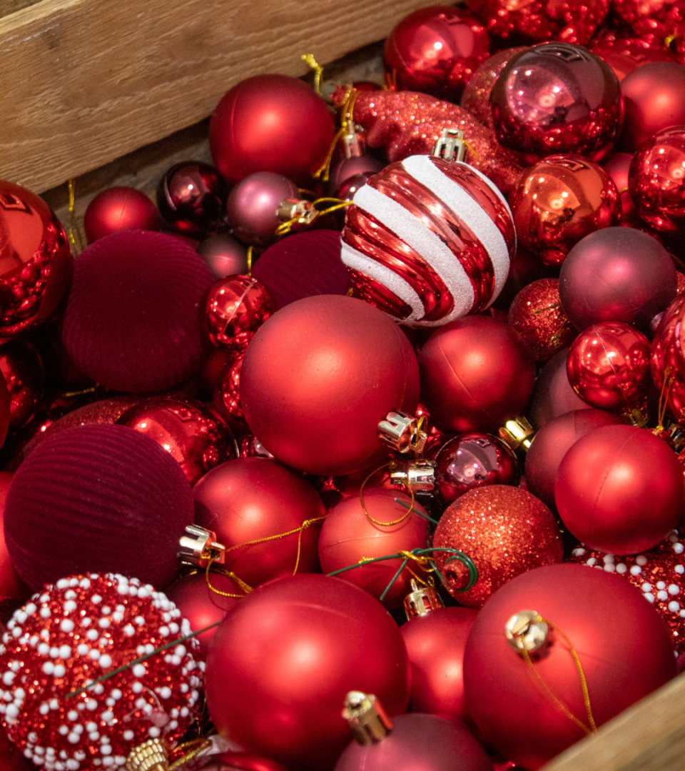 A wooden crate overflowing with an assortment of red Christmas baubles in matte, gloss, glitter, and patterned finishes, available for use in the festive arrangement workshop