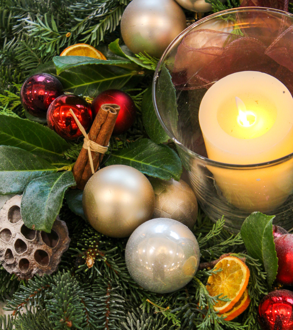 A completed Christmas table centrepiece featuring silver and red baubles, dried orange slices, cinnamon sticks, bay leaves, evergreen fir branches, a lotus seed head, and a glowing pillar candle in a glass hurricane vase