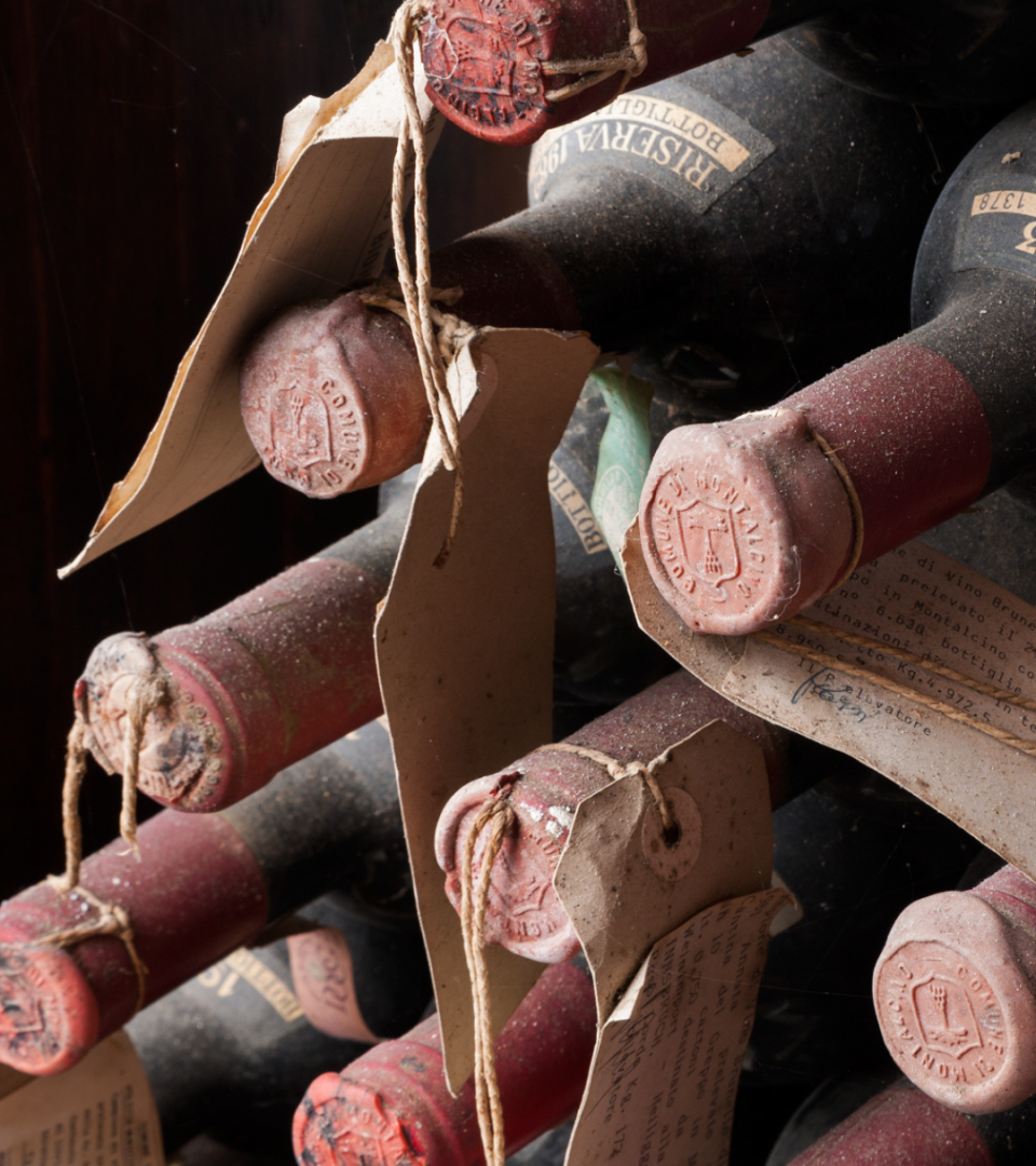 Dust-covered Biondi-Santi wine bottles stored horizontally in a cellar, with red wax seals, handwritten archival tags, and aged labels illuminated by warm cellar light.