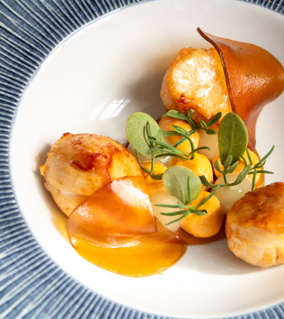 Close-up of pan-seared scallops with caramelized edges, served with baby onions, crispy sage leaves, and a glossy amber sauce, plated on a white bowl set against a blue-striped linen at Ness Walk.