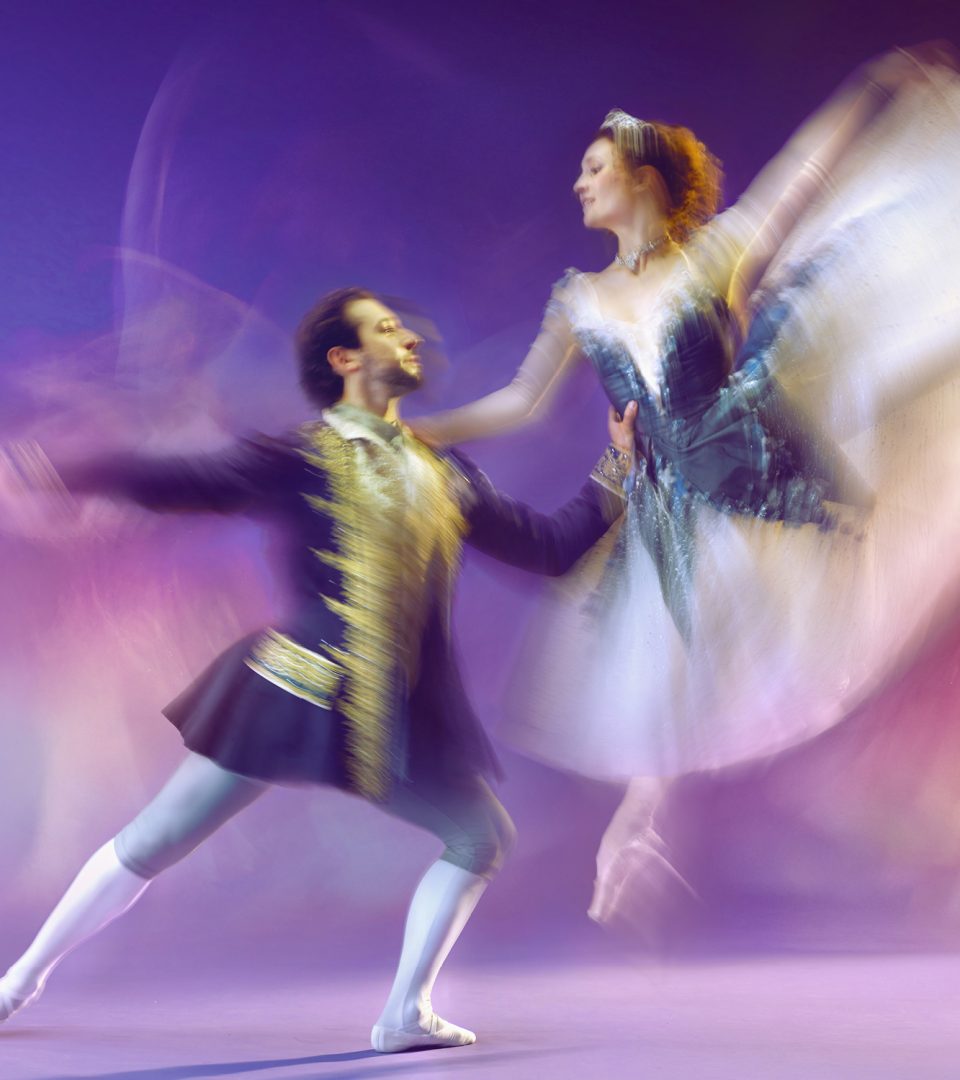 A male and female ballet dancer perform an elegant lift during a production of Cinders. Captured with intentional motion blur, the image conveys sweeping movement as the ballerina's blue-and-white gown billows dramatically and the male dancer supports her with outstretched arms. Purple, pink and gold stage lighting creates a dreamy, magical atmosphere, emphasising the energy and romance of the performance.