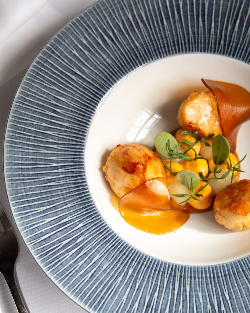 Perfectly seared scallops served during Christmas Day lunch at Ness Walk, accompanied by silky vegetable purées, rich sauce, delicate herb garnish and crisp vegetable crisps, beautifully presented in an elegant blue-rimmed bowl.