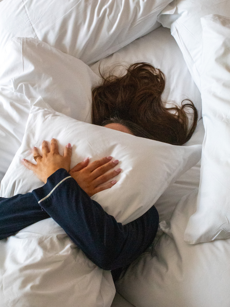 Overhead view of a person in navy pajamas hugging a white pillow while lying in bed surrounded by additional white pillows and bedding, face mostly hidden by their hair.